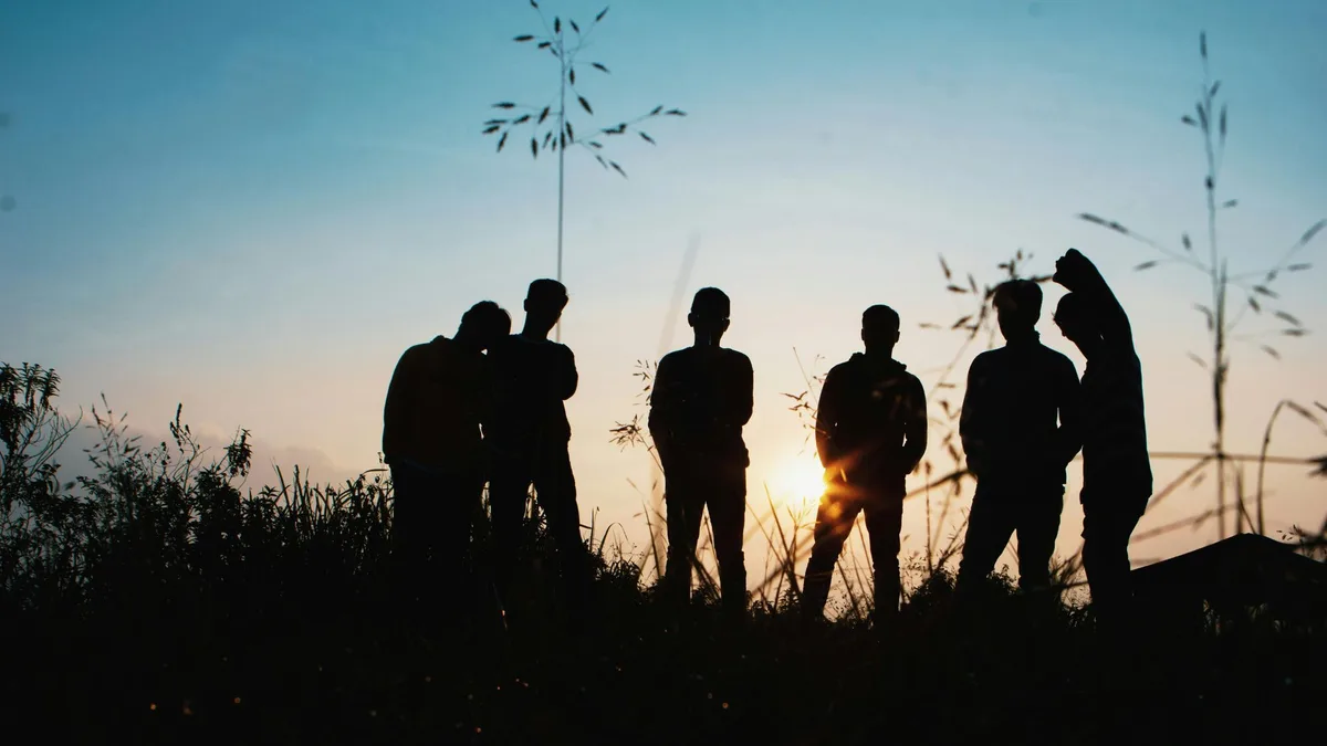 Silhouettes of young people standing together at sunset against tall grasses.